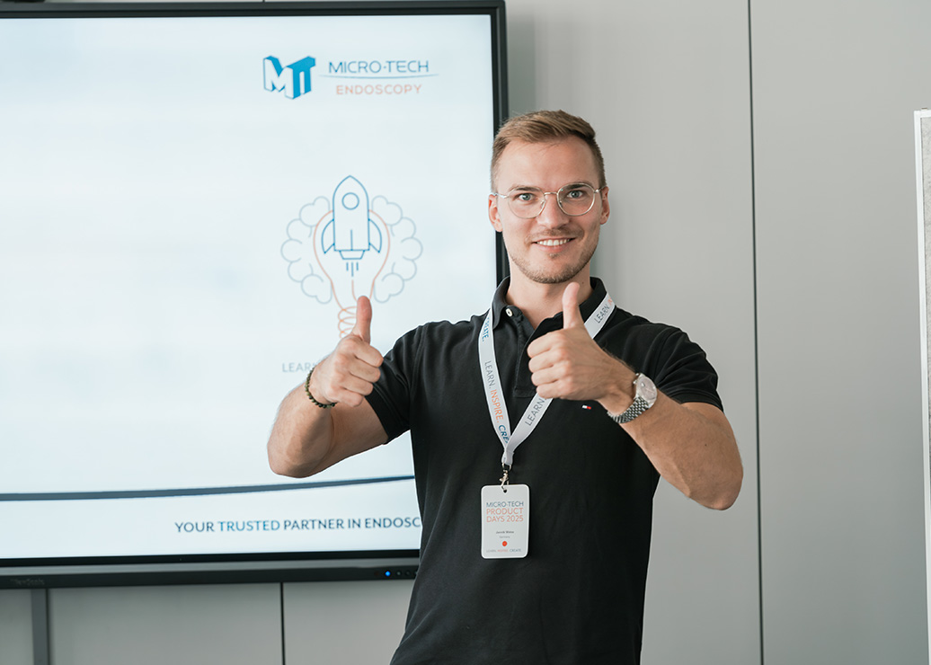Mitarbeiter bei MICRO-TECH Endoscopy steht vor Präsentationsscreen und zeigt mit beiden Händen Daumen hoch.